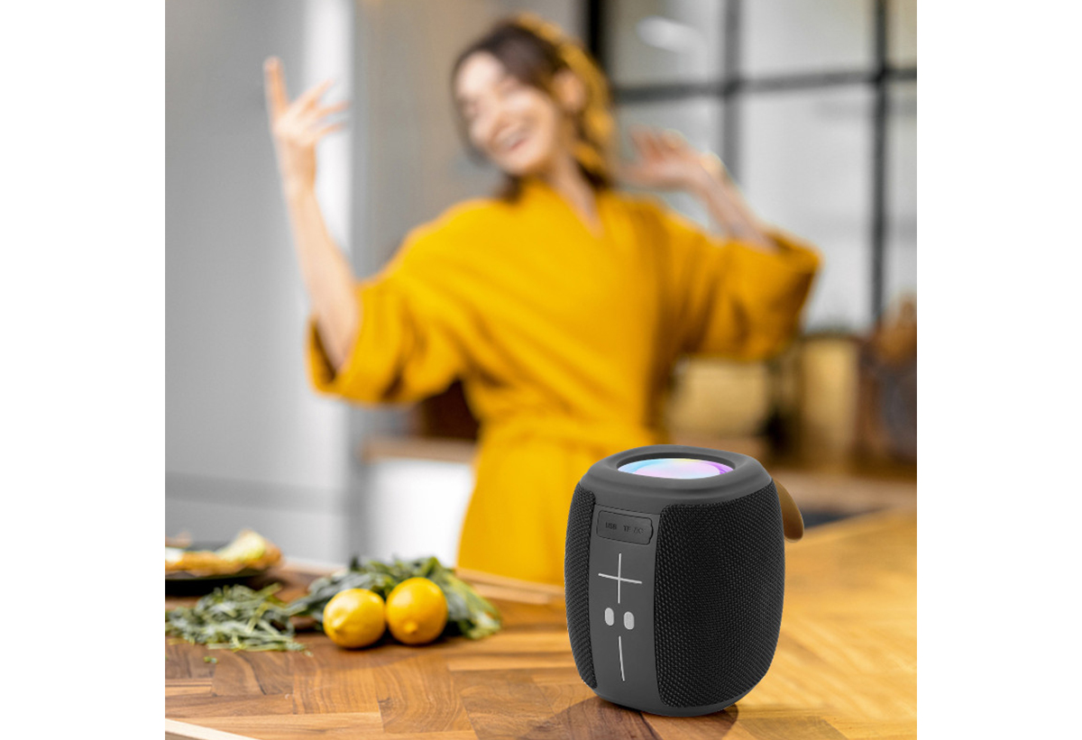 Απεικονίζεται το ηχείο σε τραπέζι  κουζίνας. Πίσω μια γυναίκα χορεύει.